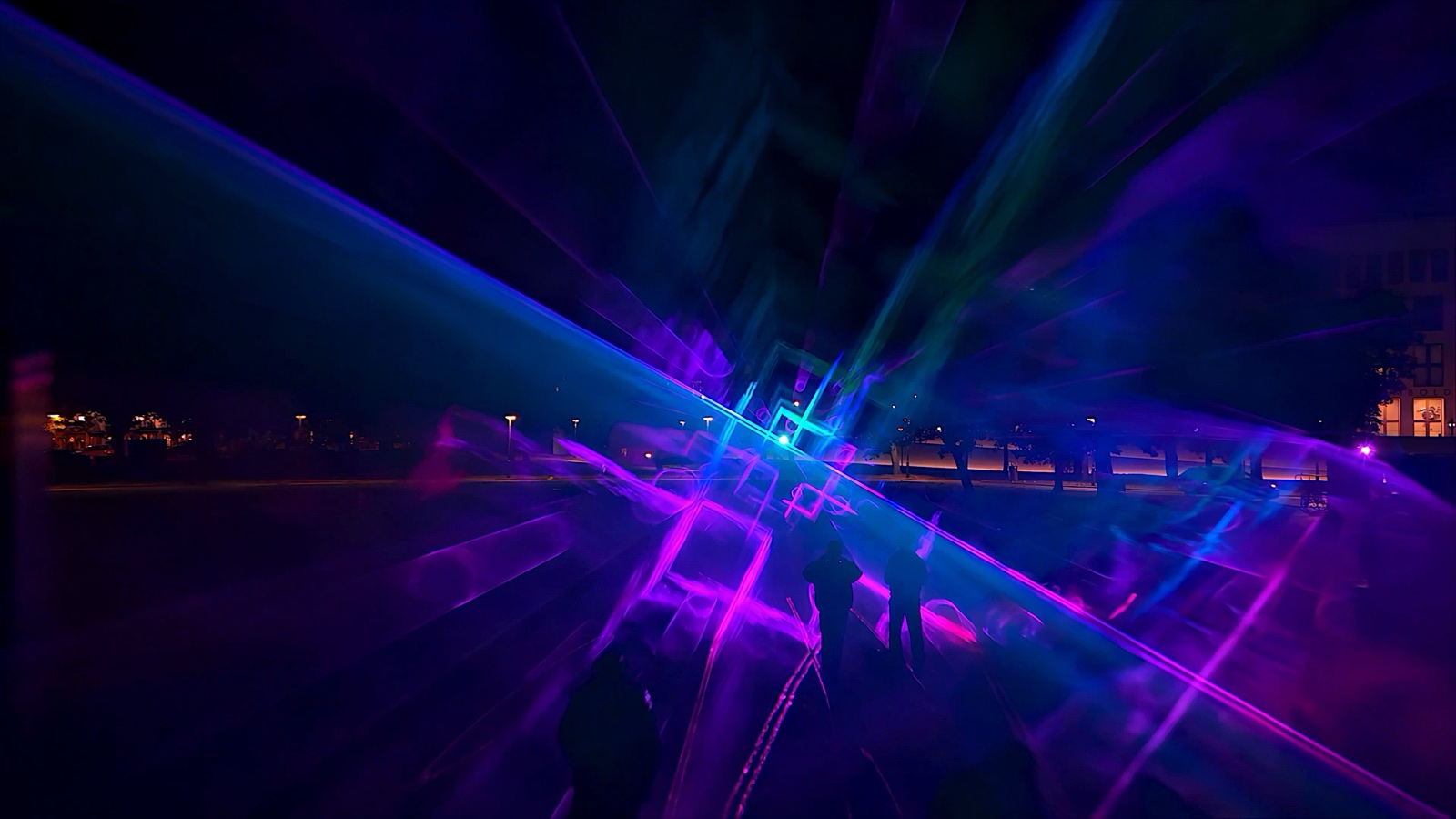 Blue and violet beams crossing low over the square, a bright cyan core at the centre and city lights beyond