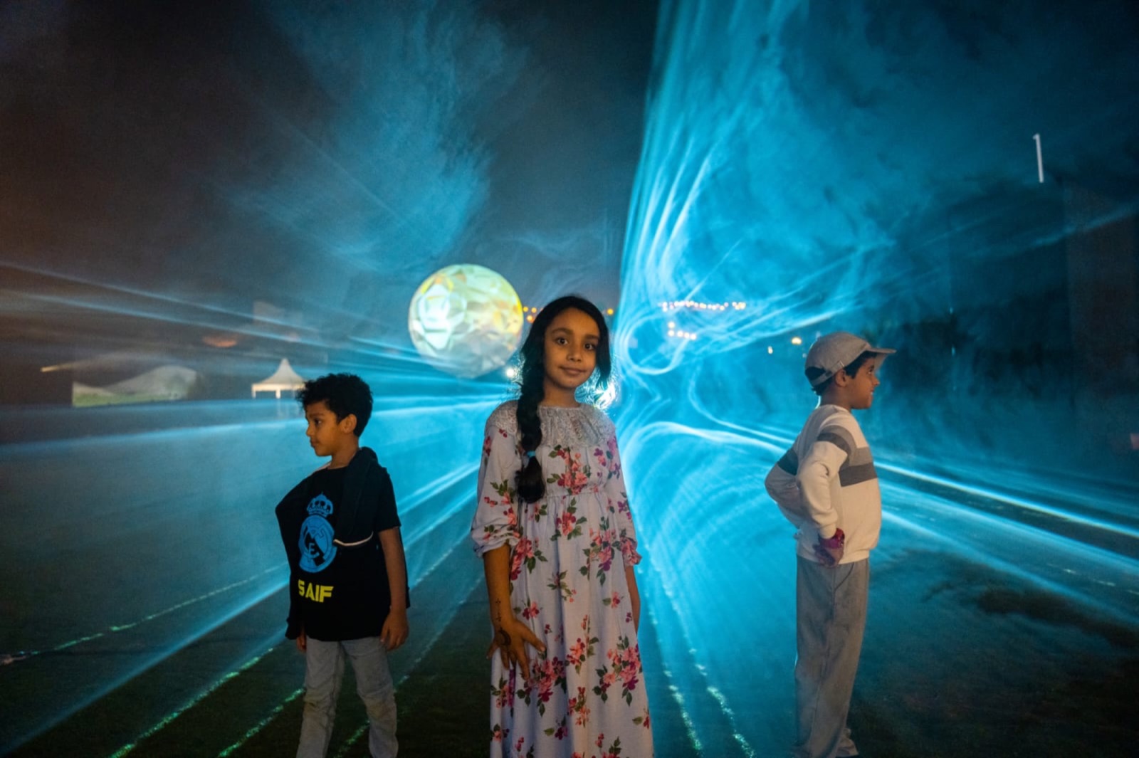 Three children standing inside sheets of blue light drawn through fog on the festival grounds, the projection sphere glowing behind them