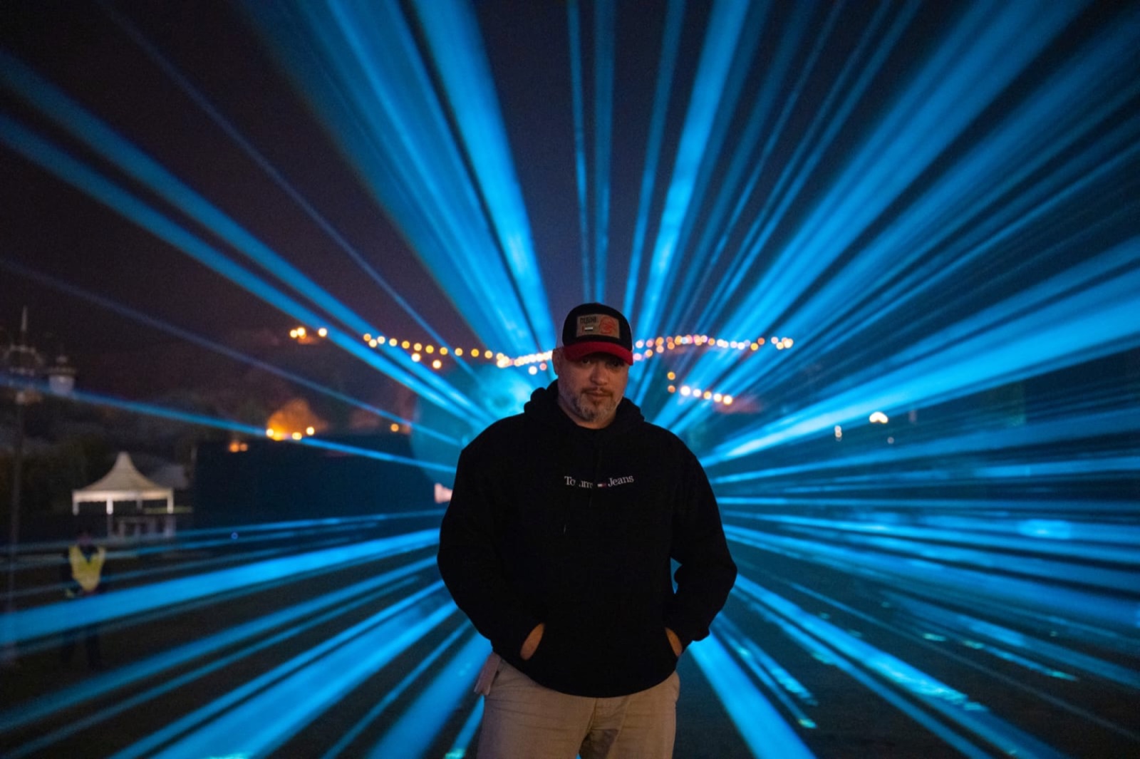 A man in a cap standing in front of a fan of blue light beams drawn through fog on the festival grounds at night