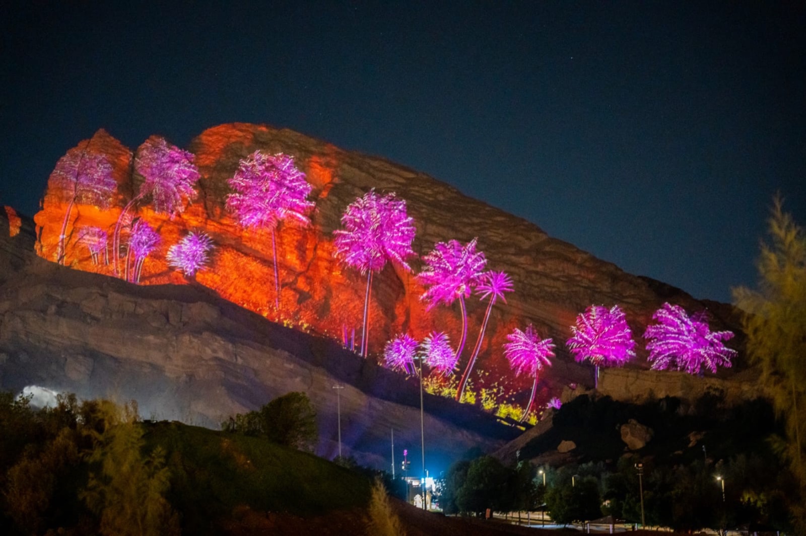 Palm trees projected in pink and violet across an orange-lit rock face at the foot of Jebel Hafeet at night, with trees in the foreground