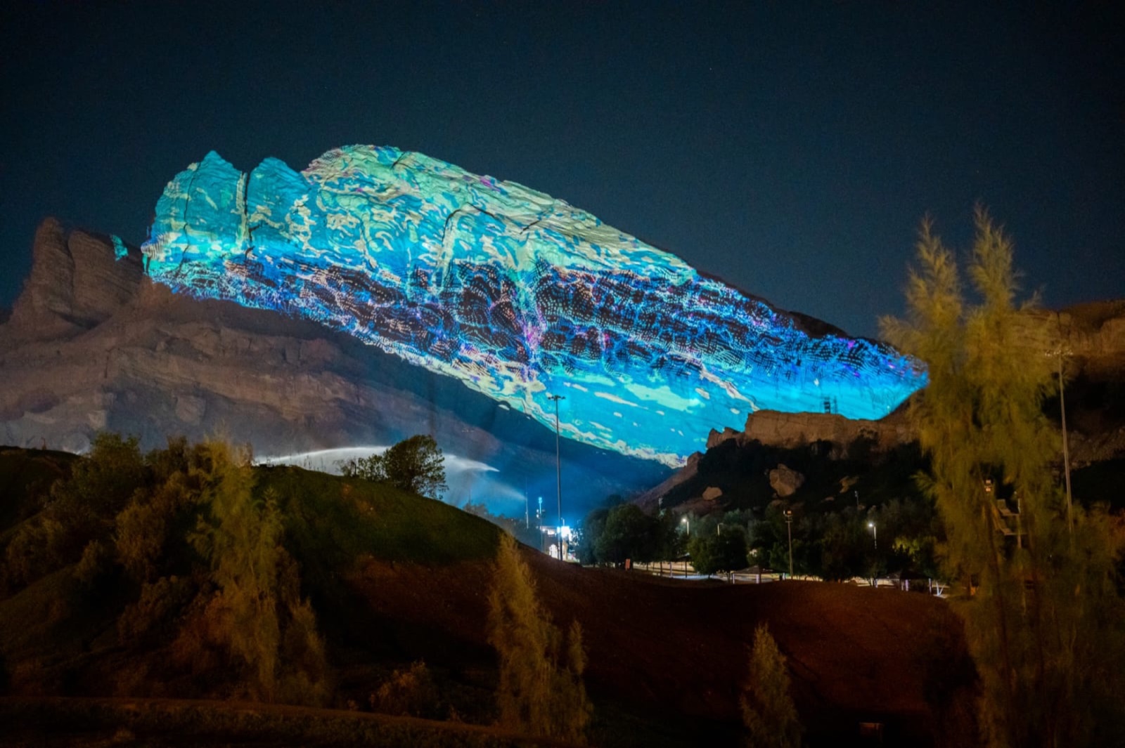 A rock face at the foot of Jebel Hafeet covered in a turquoise and blue projected texture at night, trees in the foreground
