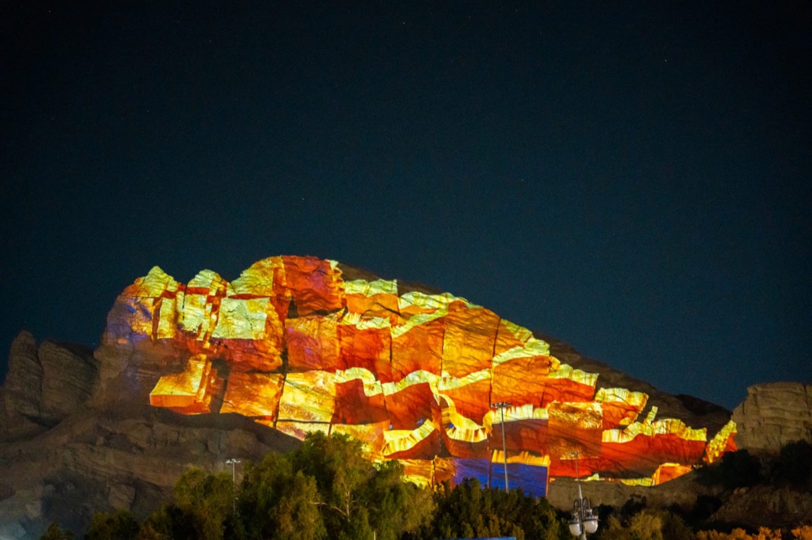 A rock face at the foot of Jebel Hafeet mapped with layered orange and yellow bands at night, above a line of trees
