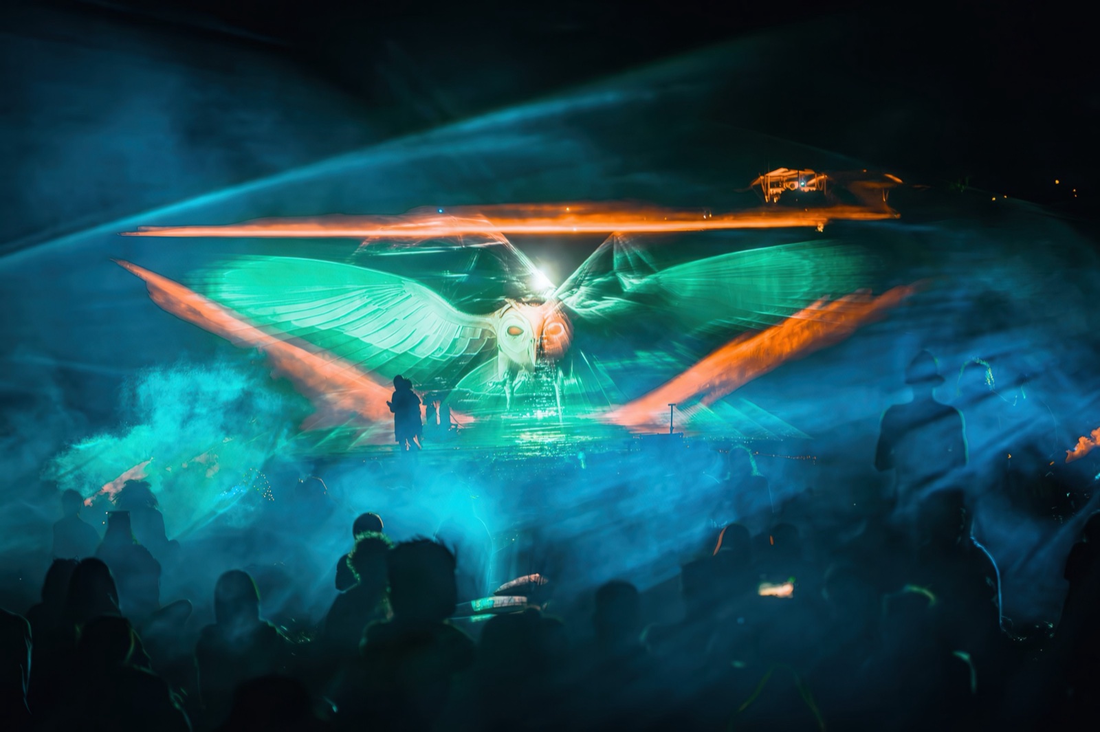 A wide blue and green light form spanning the night sky above the festival crowd, mist catching the projection beams