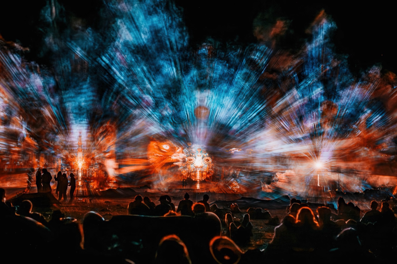 A burst of blue and orange light radiating outward above silhouetted festivalgoers, beams crossing in the haze