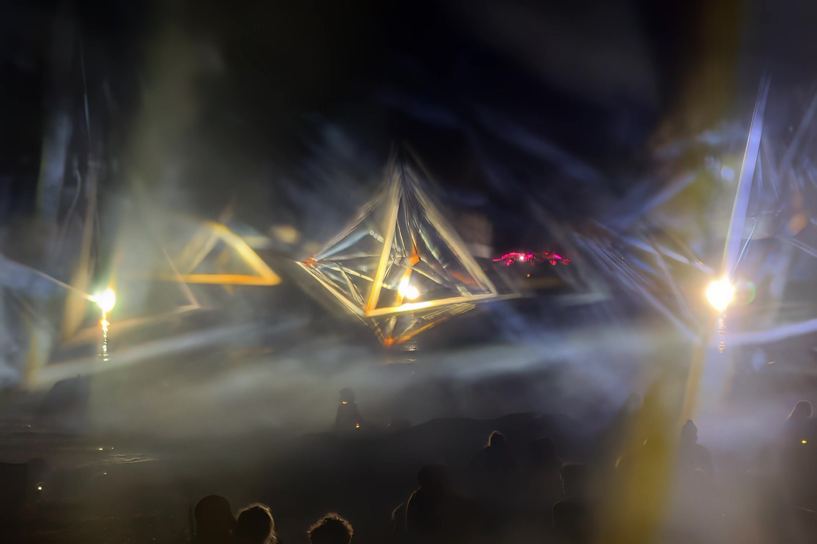 Golden pyramid-shaped outlines of light hanging in the haze above the heads of the audience, bright lamps glowing through the mist
