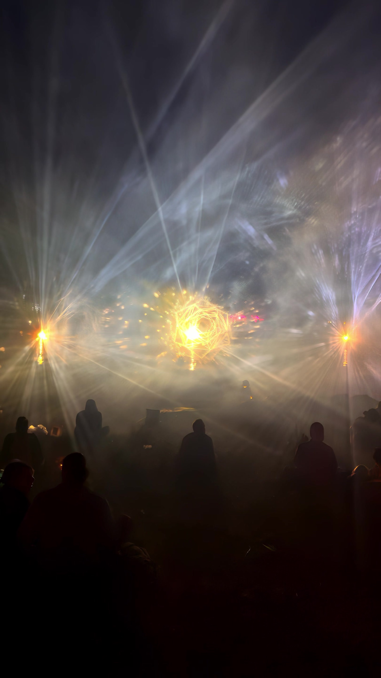 A festival crowd at night in front of BEYOND at Boom Festival: a glowing golden form of light hangs in drifting fog above the lake while beams fan out over the audience