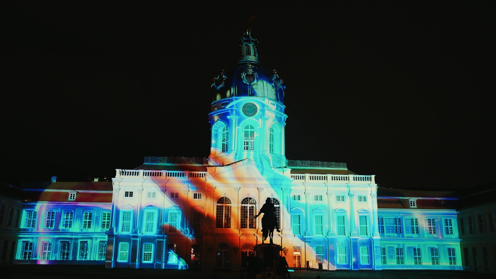 Charlottenburg Palace at night in turquoise and blue projected light, a giant orange hand sweeping across the centre and the equestrian statue in silhouette — The Cathedral of the Infinite Scroll by RE:SORB, Festival of Lights Berlin 2025