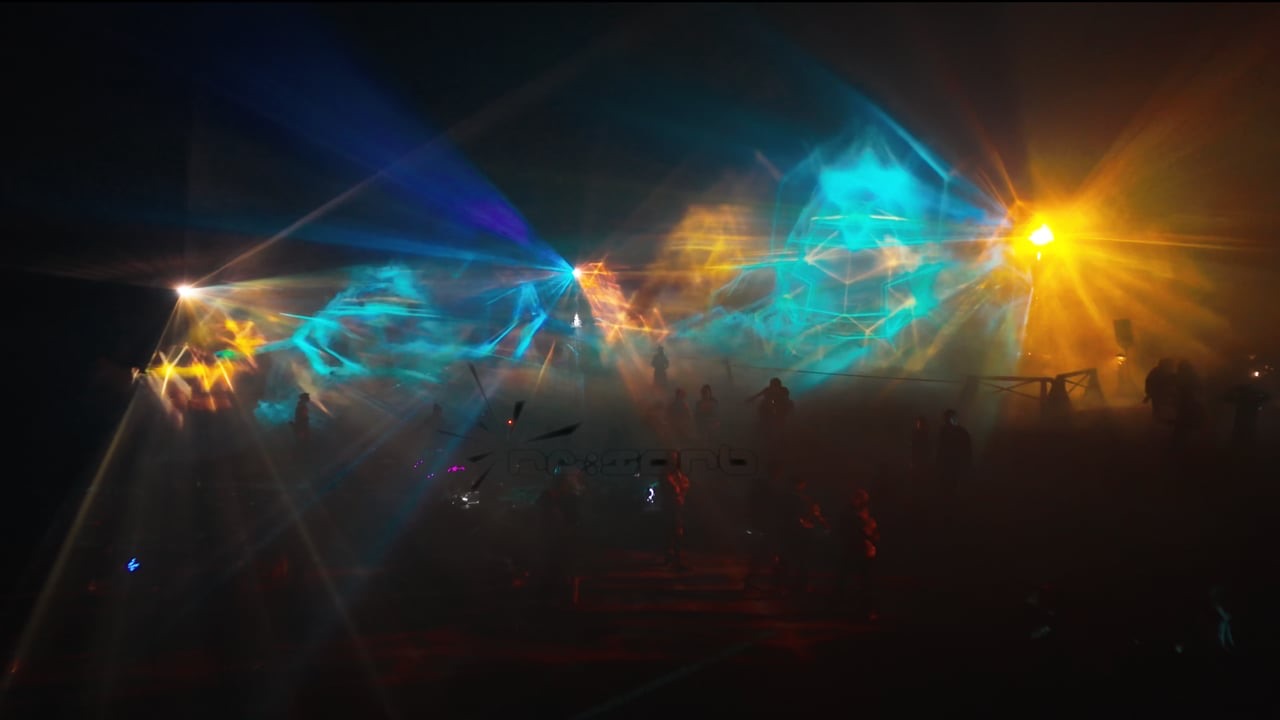 Beams of blue and orange light passing through layered water and mist screens at night, with festival visitors silhouetted in front of the Depth/100 installation at Ozora