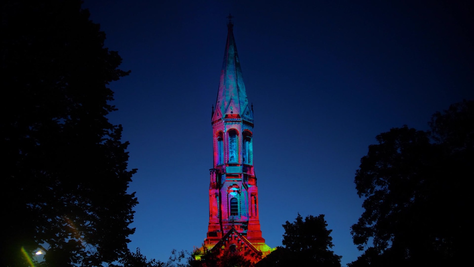 The tower of the Emmauskirche in Berlin-Kreuzberg at dusk, lit red, pink and turquoise by a projection mapping and framed by trees
