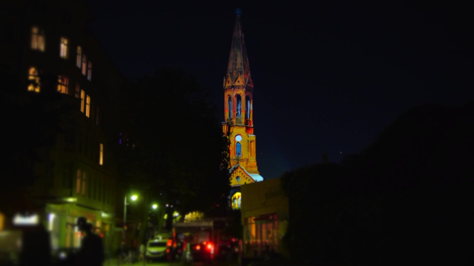 The projected church tower glowing orange above a Kreuzberg street at night, with lit windows, street lamps and passers-by