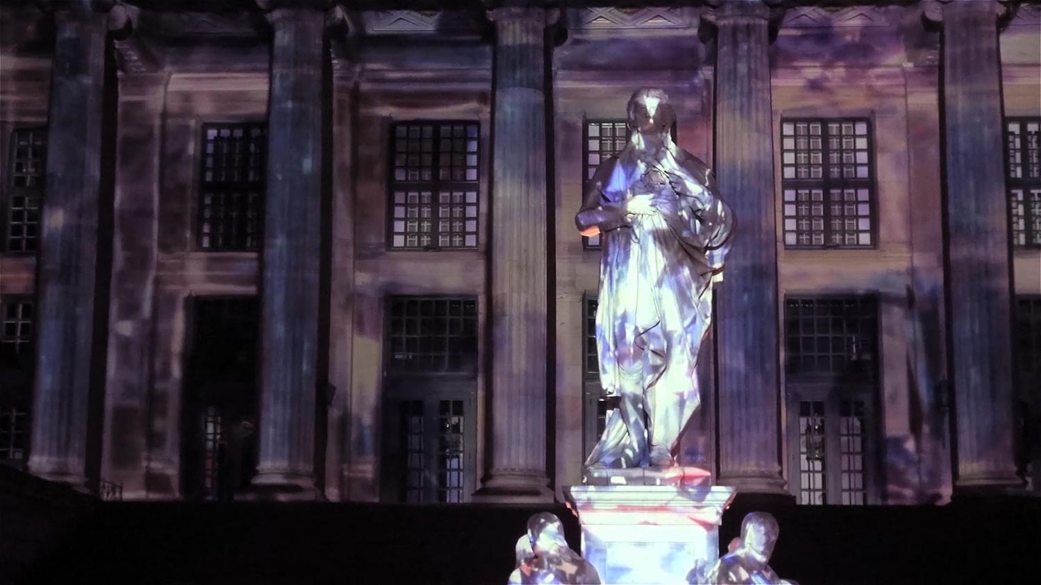 The columned façade of the Konzerthaus at Gendarmenmarkt in Berlin lit by a colourful projection mapping with the Schiller monument in front