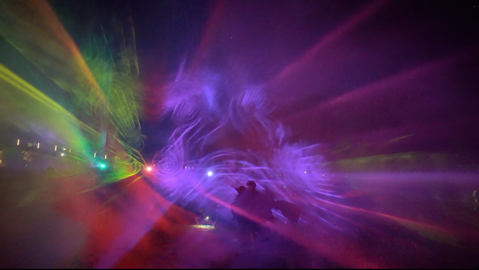 A crowd at night in front of a mapped building at Boom Festival, with beams of blue and white light passing through fog in front of the façade