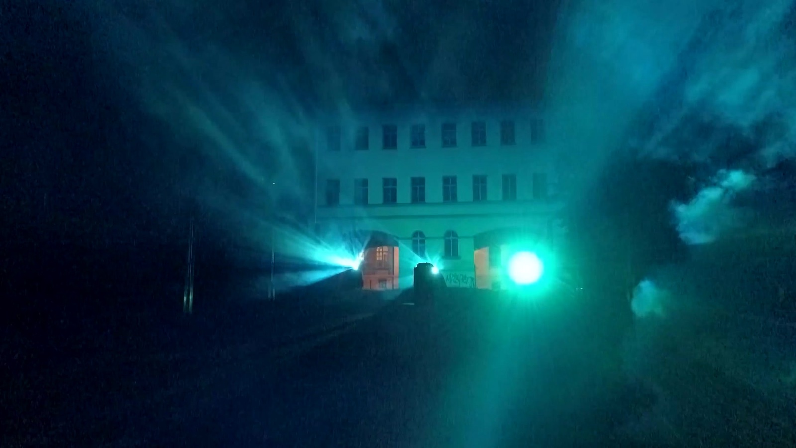 A crowd at night in front of a mapped building in Weimar, with beams of blue and white light passing through fog in front of the façade