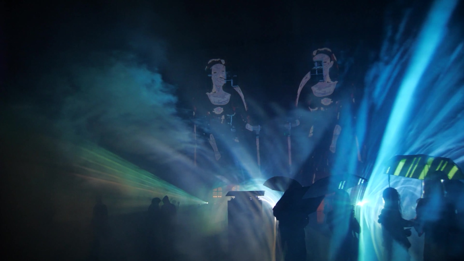 Two tall ornamental figures either side of the stage, blue beams and smoke rising between them