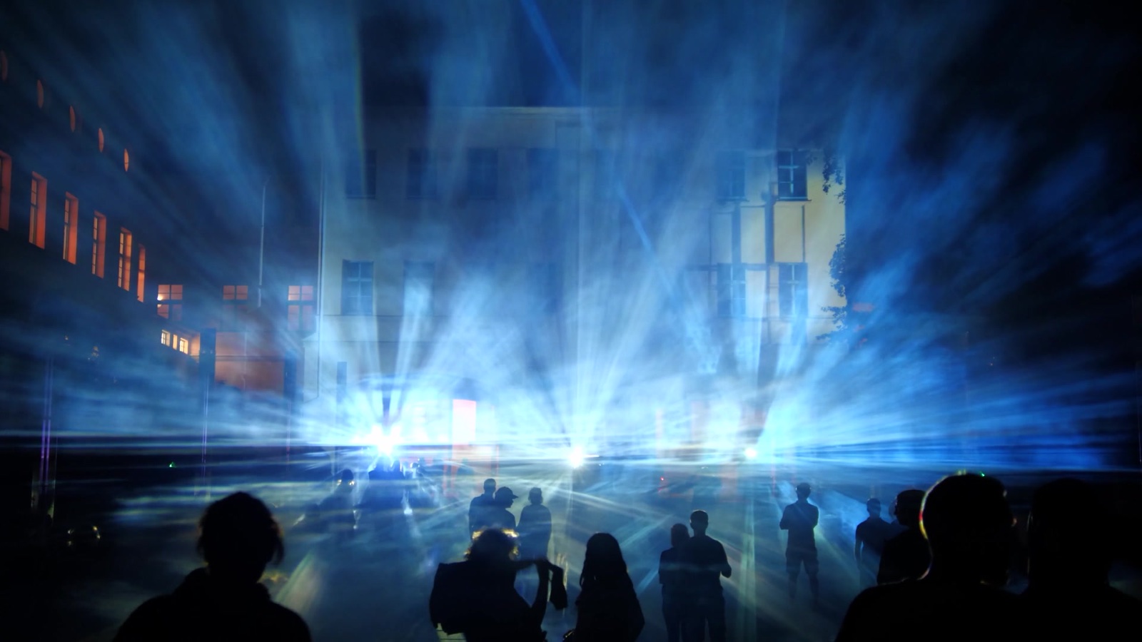 A bright blue-white burst of light in front of a building, spectators standing in silhouette along the foreground