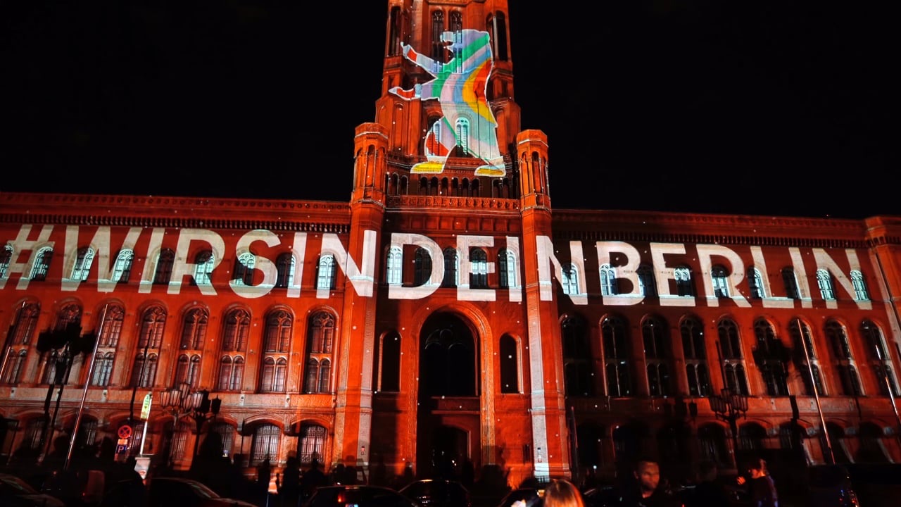 Das Rote Rathaus in Berlin bei Nacht: ein regenbogenfarbener Berliner Bär auf dem Turm und #WIRSINDEINBERLIN in weißen Großbuchstaben über dem obersten Geschoss der Backsteinfassade, im Vordergrund Zuschauer und parkende Autos
