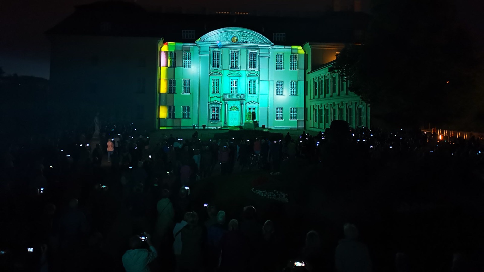 Schloss Köpenick lit turquoise by the projection mapping, with coloured light along the edges of the façade and the crowd in the foreground