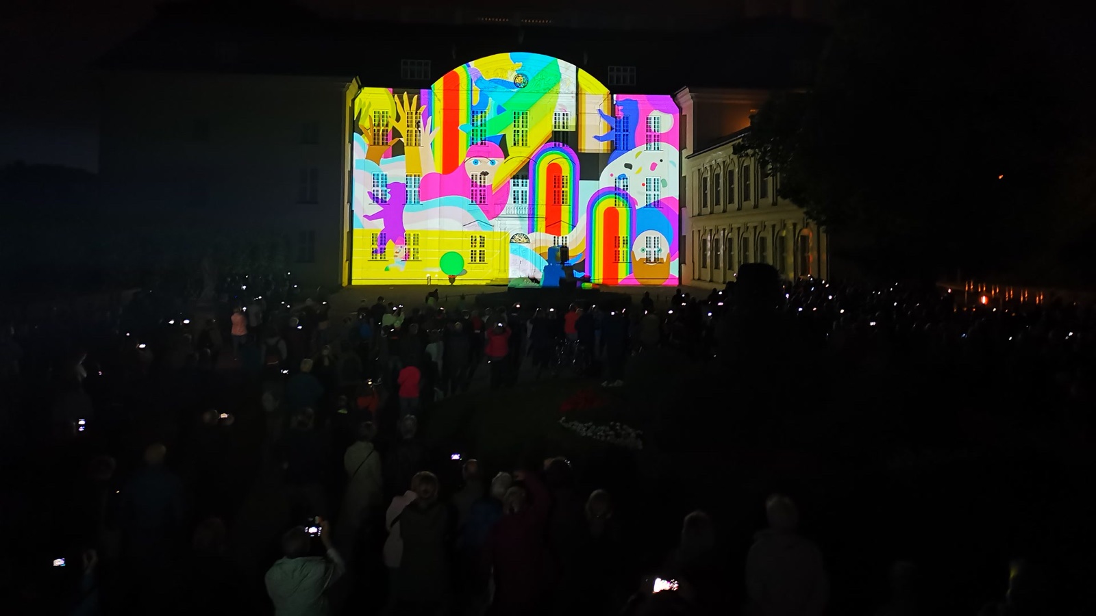 A colourful illustrated motif of rainbows and houses projected onto the central façade of Schloss Köpenick, the audience in silhouette