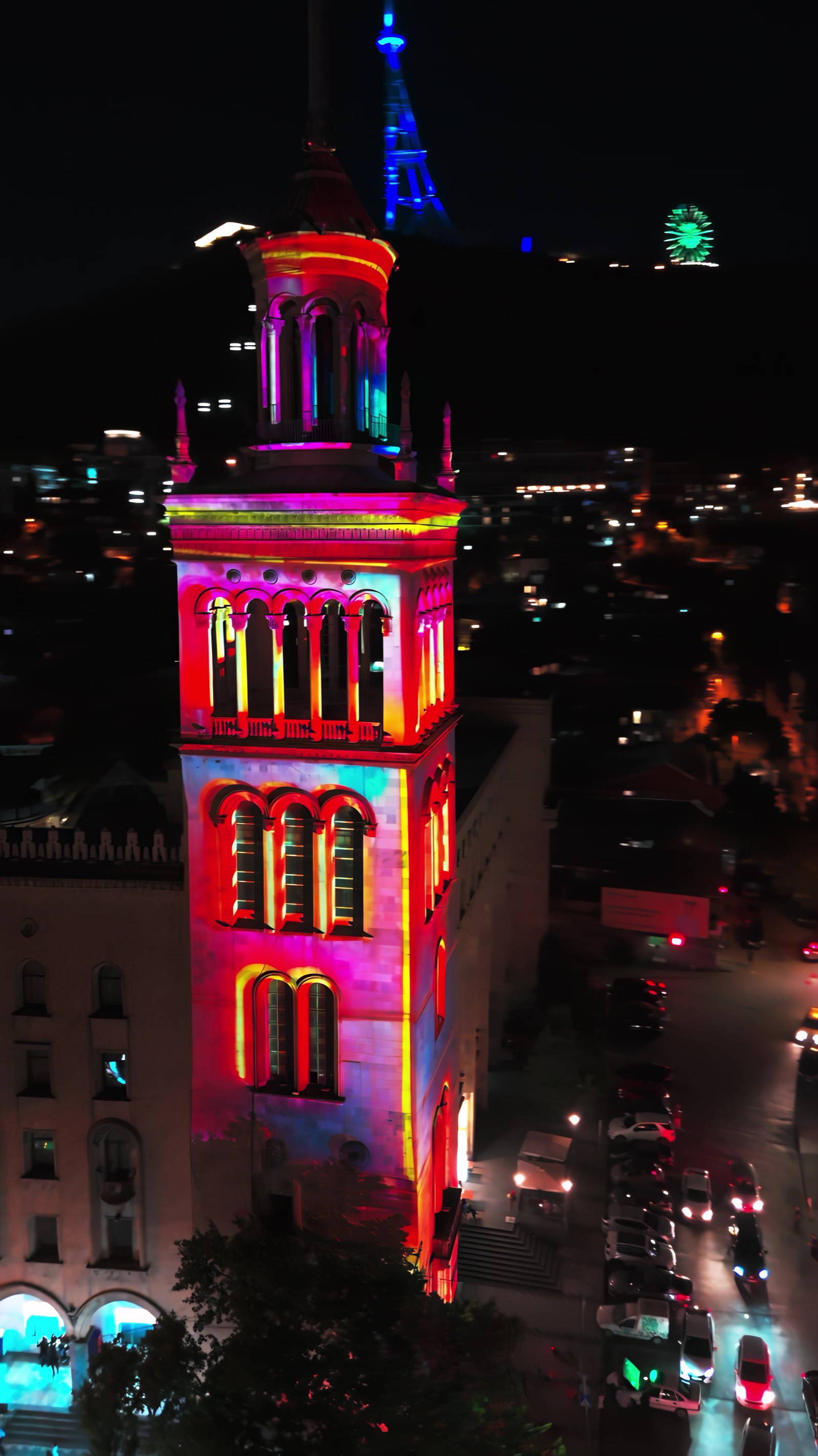The tower of the Georgian National Academy of Sciences on Rustaveli Avenue in Tbilisi lit in pink and red by RE:SORB’s projection mapping at night, Mount Mtatsminda with its TV tower and Ferris wheel behind
