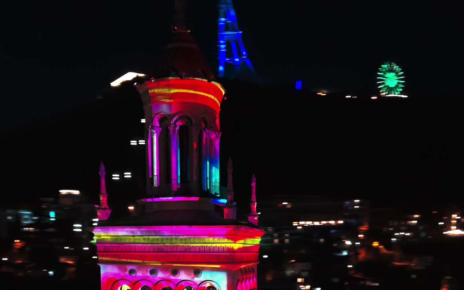 The crown of the Academy of Sciences tower in Tbilisi lit in pink and red by projection mapping, the Mtatsminda TV tower and Ferris wheel glowing on the hill behind