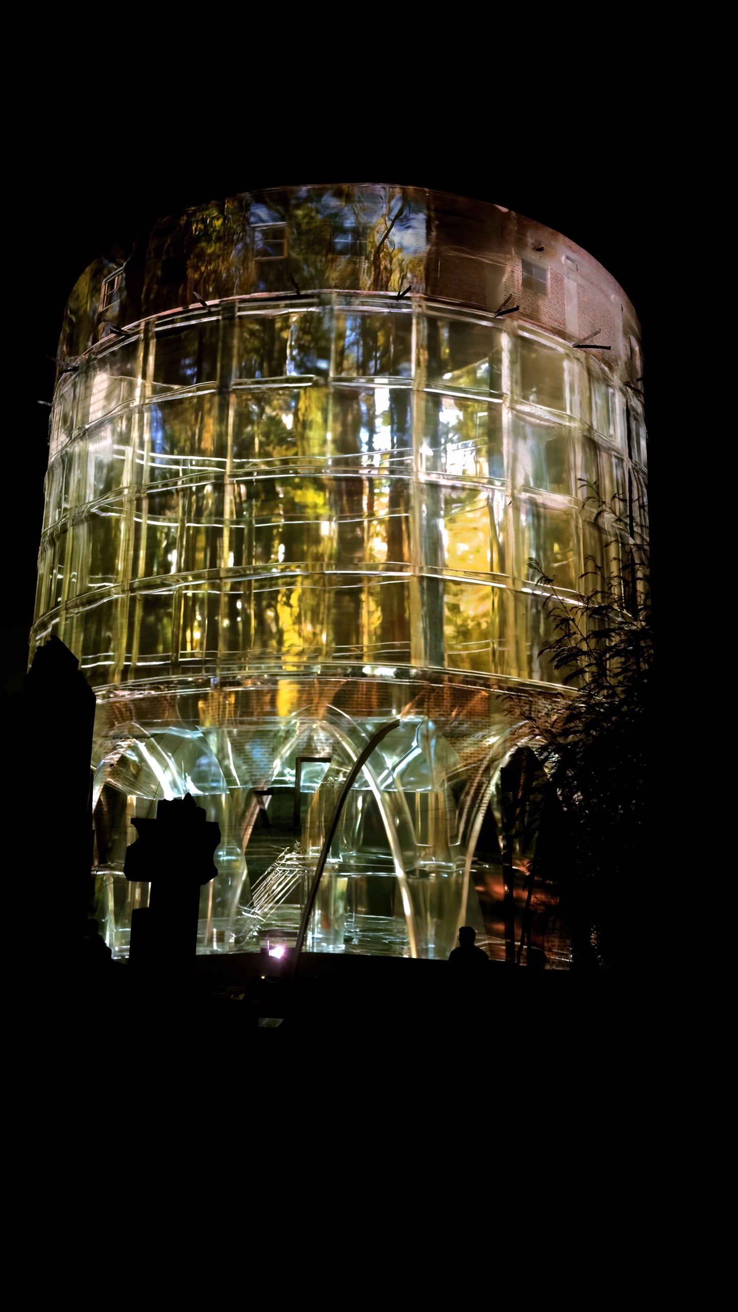 The silo lit in gold and amber, its projected lattice of ribs and beams standing out against the night while visitors watch from the ground