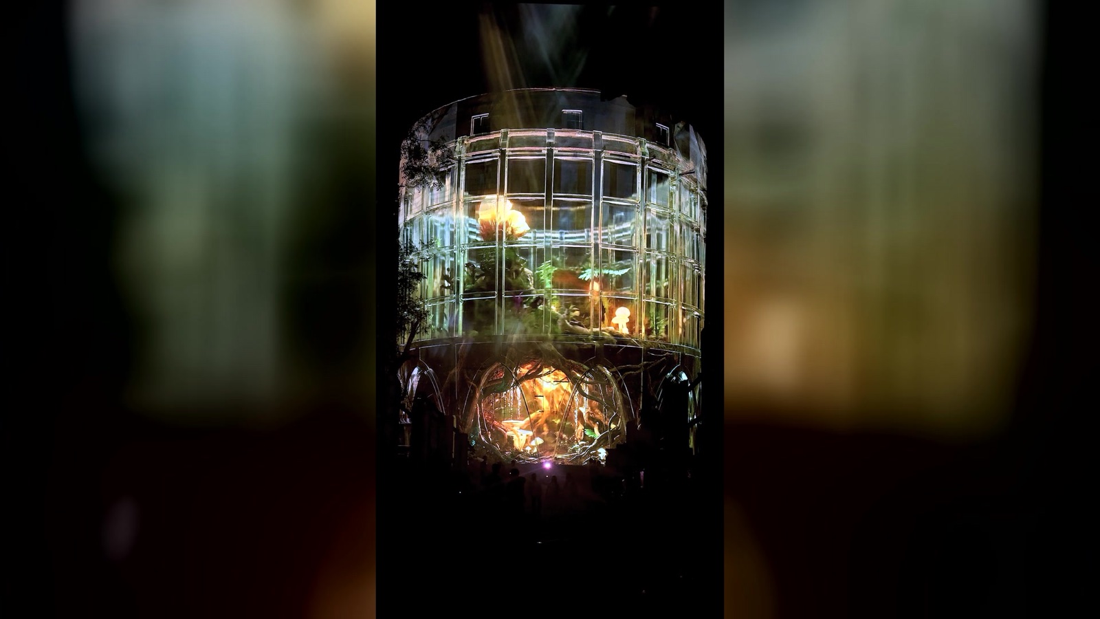The concrete silo of the Alembic Glass Factory in Bengaluru rendered as a transparent lattice by projection, with an amber interior glowing inside and holographic fog around it