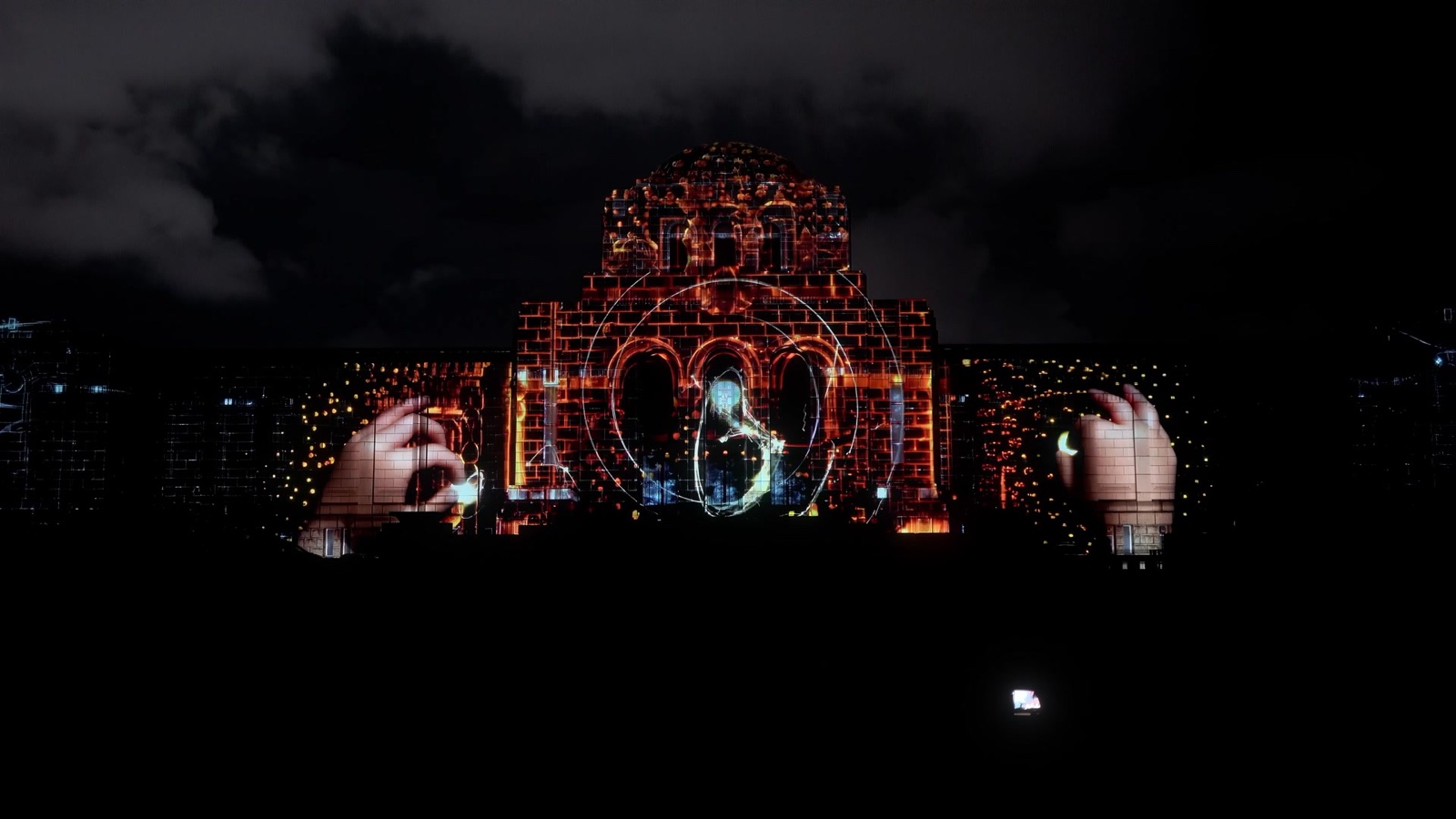 A façade in Tokyo lit in red and orange by a projection mapping in which two giant hands reach in from either side towards a figure drawn in light inside a circle